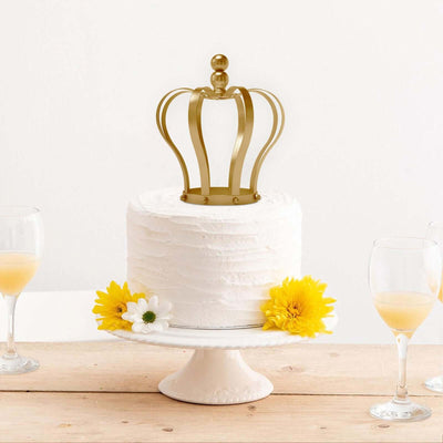White frosted cake topped with a gold crown, surrounded by yellow flowers and drinks.