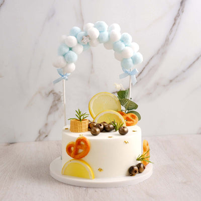 Decorative cake topped with fruit and a colorful arch of pom-poms.