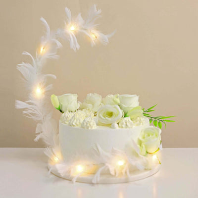 White floral cake with decorative lights and feathers.