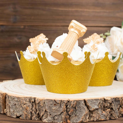 Gold crown cupcake wrappers with decorative toppers on a rustic wooden slice.