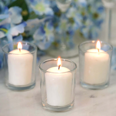 Three white candles in glass holders with blue flowers in the background.