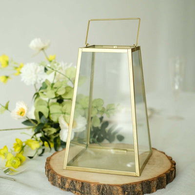 Glass lantern with gold accents on wooden slice, surrounded by flowers.