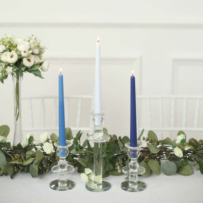 Three elegant candles in glass holders on a table with greenery and flowers.