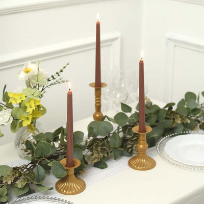 Elegant gold candle holders with brown candles and greenery on a dining table.