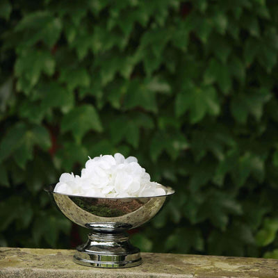 Silver bowl filled with white flowers on a stone surface surrounded by greenery.
