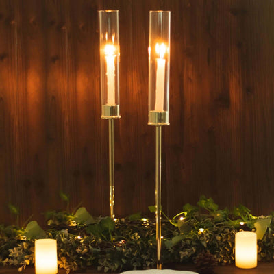 Elegant candle holders with glass cylinders and lit candles, surrounded by greenery.