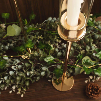 Elegant gold candle holder with glass shade and greenery backdrop.