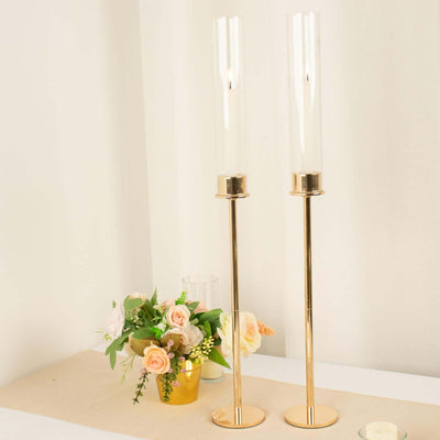 Elegant gold candle holders with glass tubes and floral arrangement in the background.