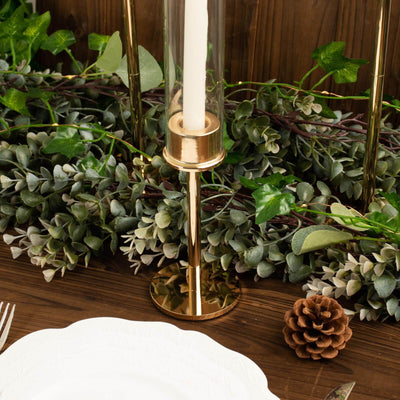 Gold candle holder with a clear glass cylinder and greenery background.