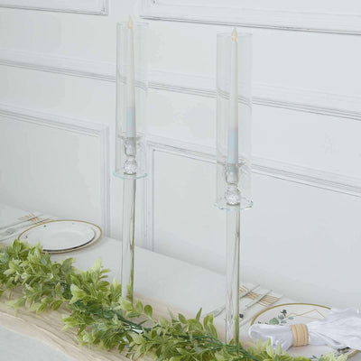 Elegant glass candle holders with candles and greenery on a table.