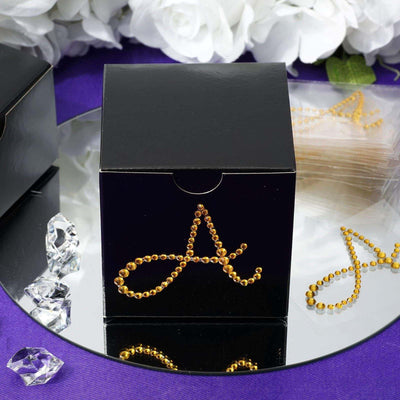 Black gift box with gold rhinestone letter "A" on a mirrored surface.