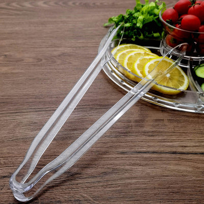 Clear acrylic serving tongs on a wooden table with fresh vegetables and fruits.