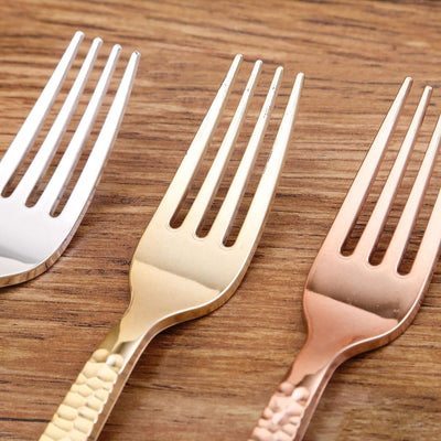 Three decorative forks in silver, gold, and rose gold on a wooden surface.