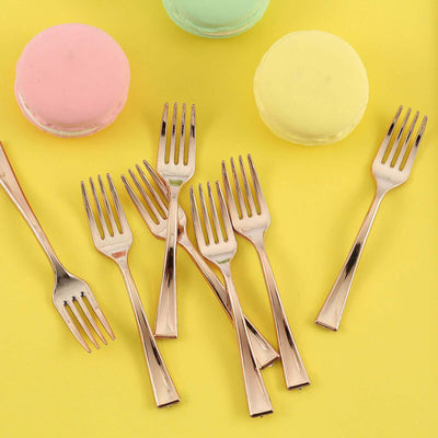 Rose gold dessert forks surrounded by colorful macarons on a yellow background.