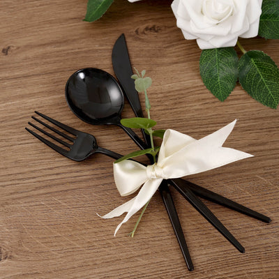 Black cutlery set with ribbon and greenery on wooden table.