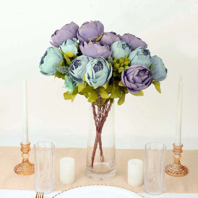 Artificial peony flower bouquet in a clear vase with candles on a table.