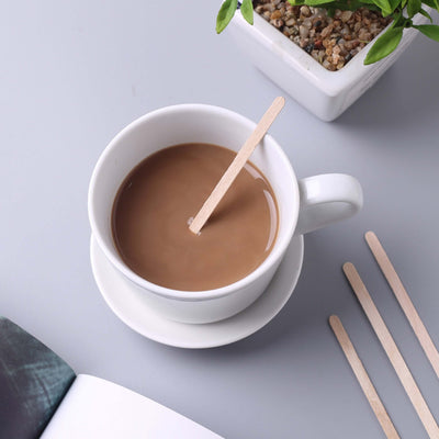 Cup of coffee with a wooden stirrer on a saucer, surrounded by a plant and book.