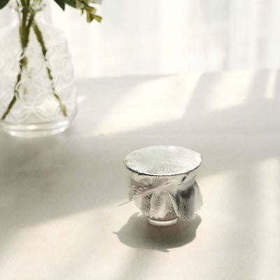 Small glass jar with a silver lid and decorative ribbon on a light surface.