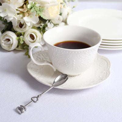 White tea cup with saucer and silver spoon, surrounded by flowers.