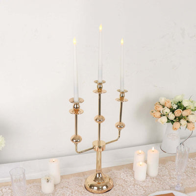 Gold candelabra with three white candles on a table with decorative flowers.