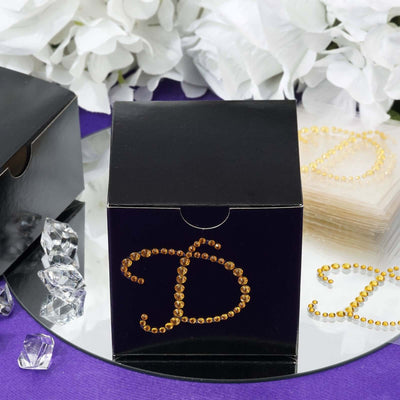 Black gift box with rhinestone letter D, surrounded by flowers and crystals.
