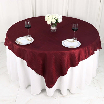 Round burgundy tablecloth draped over a white table with plates and wine glasses.