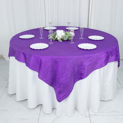Purple round tablecloth draped over a white table with plates and decorations.