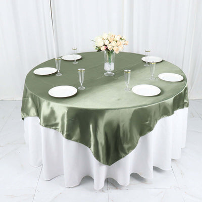 Round olive green satin tablecloth on a dining table set with plates and glasses.