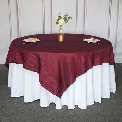 Round burgundy tablecloth on a white table with decorative vase and plates.