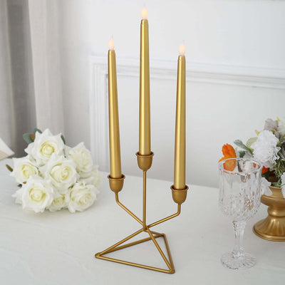 Gold three-candle holder with tapered candles and floral decor on a table.
