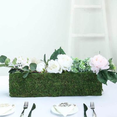 Artificial floral arrangement in a moss-covered box on a table.