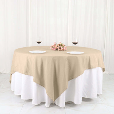 Beige round tablecloth draped over a white table with pink flowers and wine glasses.