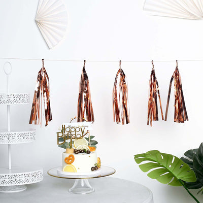 Rose gold party tassels hanging above a decorated cake on a stand.