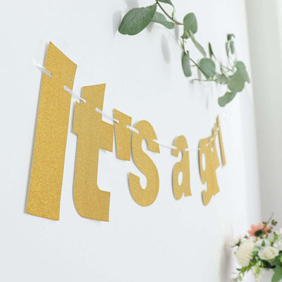 Gold "It's a girl" banner on a wall with greenery and flowers.