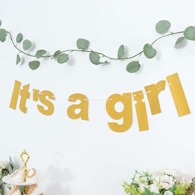 Gold "It's a Girl" banner with greenery in a festive setting.