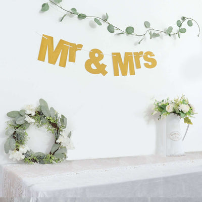 Gold "Mr & Mrs" banner with greenery and floral decor on a white backdrop.