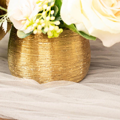 Gold textured vase with flowers on soft fabric backdrop.