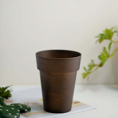 Bronze flower pot on a table with greenery in the background.