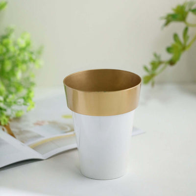 White pot with gold rim on a table with greenery in the background.