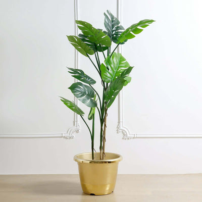 Artificial monstera plant in a gold pot against a plain background.