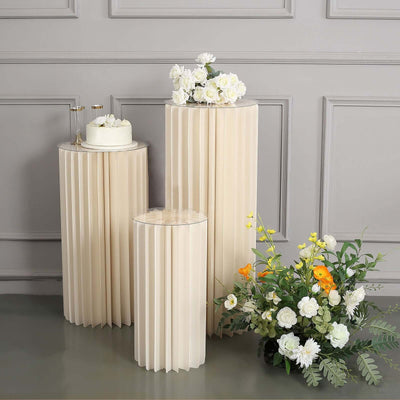 Three elegant cream-colored pleated pedestals with floral arrangements and a cake.
