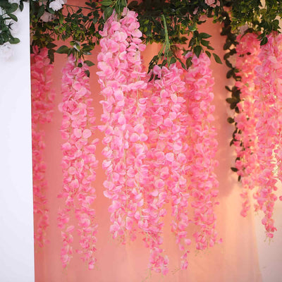Pink artificial wisteria flowers hanging from greenery backdrop.