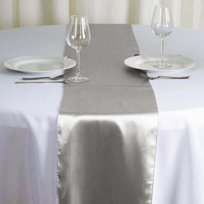 Silver satin table runner on a white tablecloth with dinnerware set.