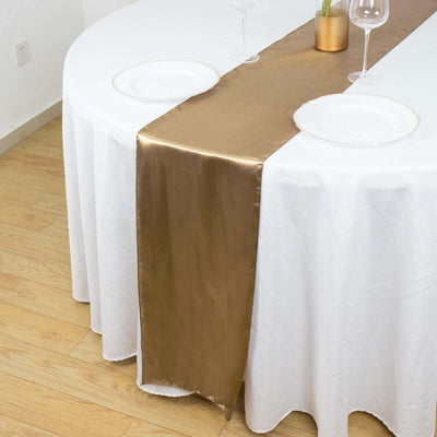 Gold satin table runner on a round table with white tablecloth and glassware.