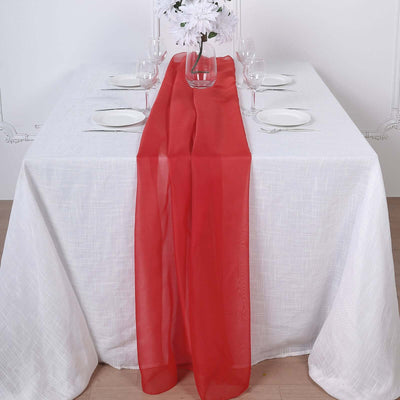 Red sheer table runner on a white table setting with glassware and plates.