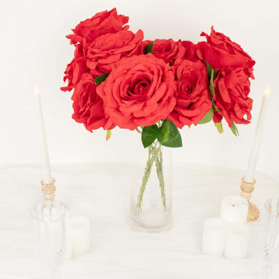 Red artificial roses in a clear vase with candles on a white table.