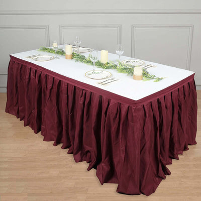 Elegant dining table set with burgundy skirt, plates, candles, and greenery centerpiece.