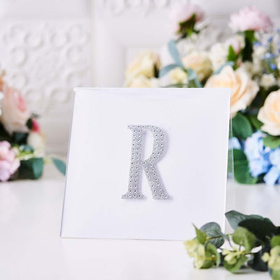 Silver glitter letter "R" on a clear display stand with floral background.