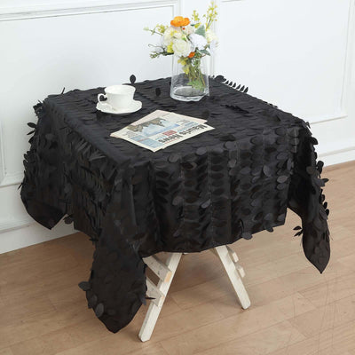 Black leaf-pattern tablecloth draped over white side table with flowers and cup.