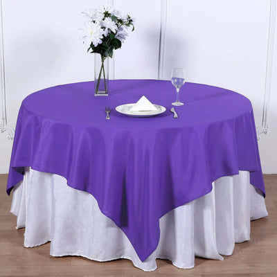 Purple round tablecloth draped over a white table with a glass and vase.
